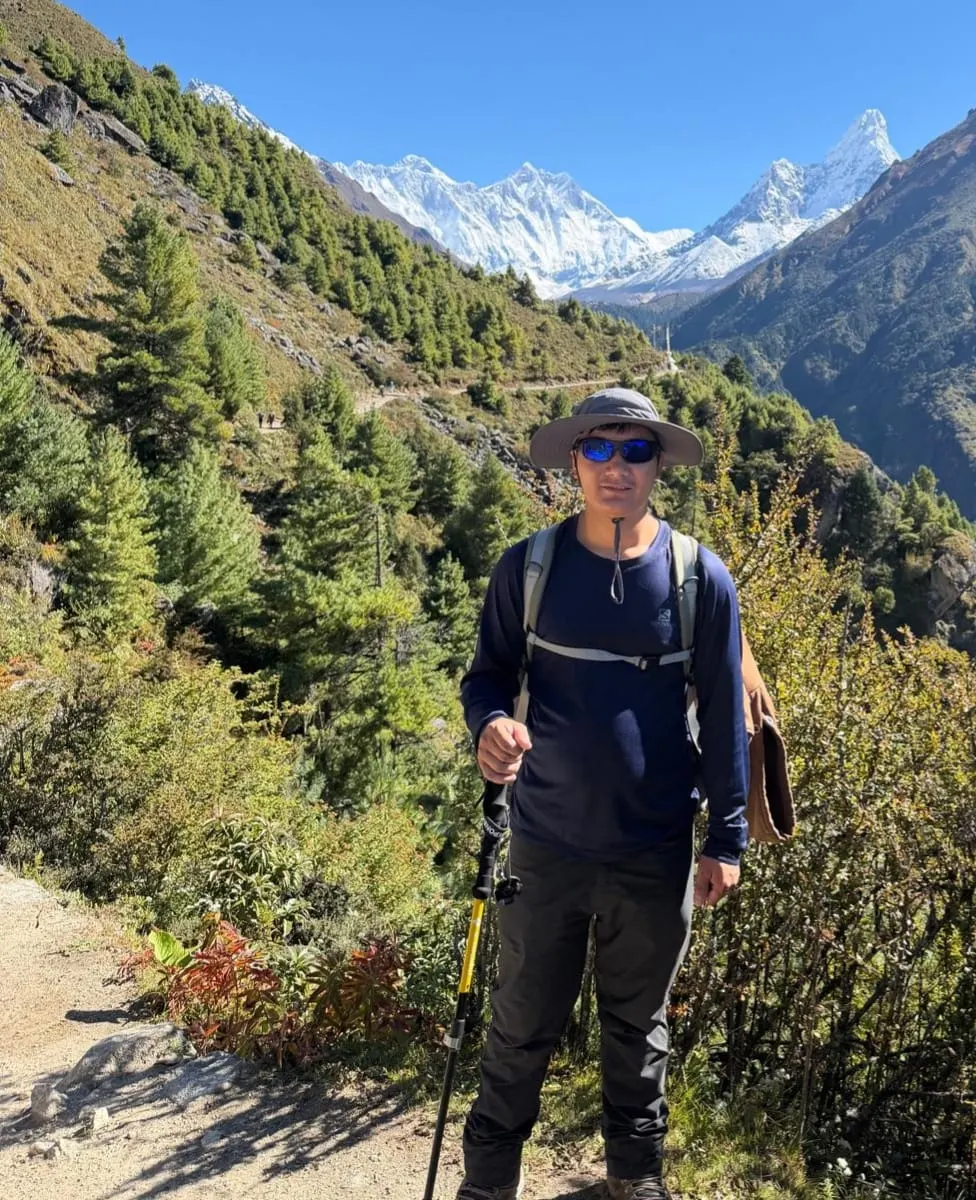 Dawa Sherpa with a backpack and trekking pole on a forest trail, snow-capped peaks behind him