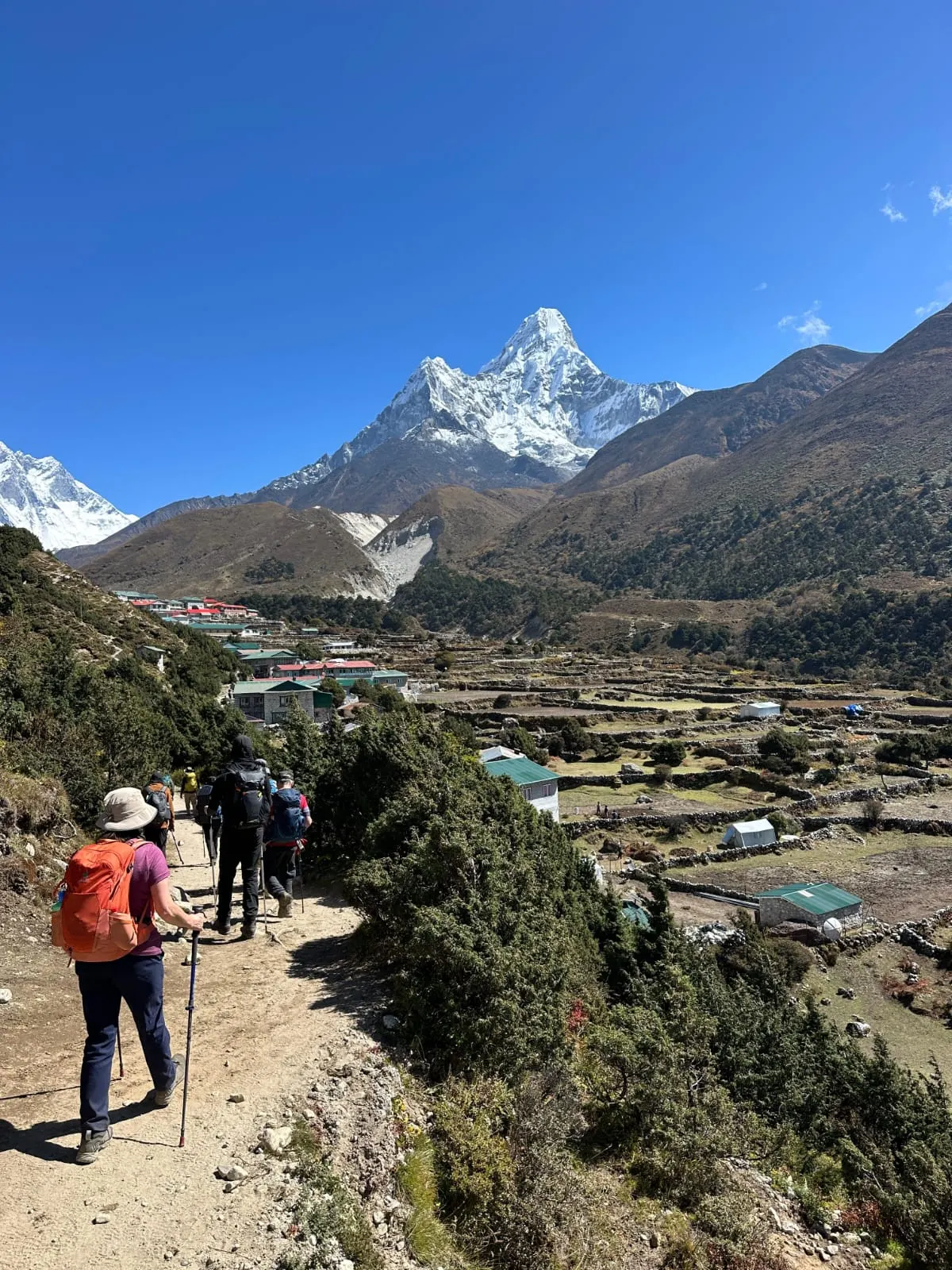 A trekker with poles on a dirt trail above a village, a sharp snow peak ahead